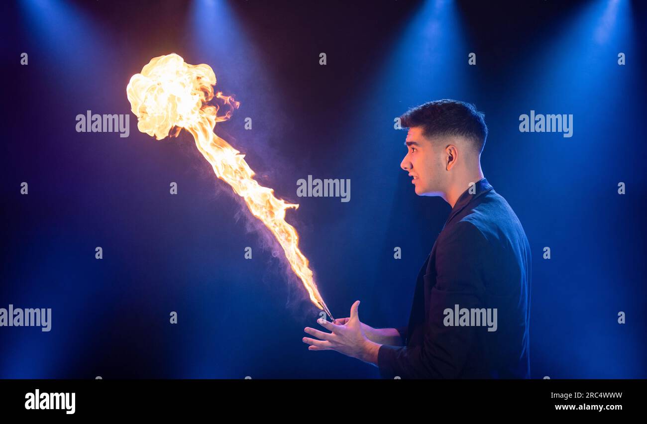 Vue latérale du prestidigitateur masculin en costume noir montrant le tour de magie avec la flamme brûlante jouant sur la scène sombre dans le cirque Banque D'Images