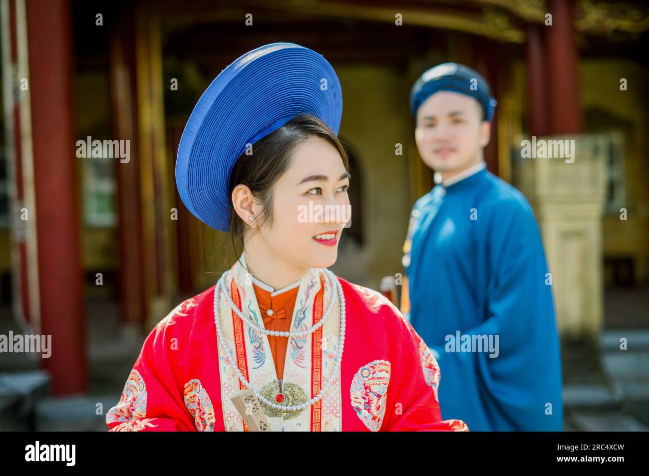 Portrait de jeune femme asiatique positive dans des vêtements traditionnels et des coiffures regardant loin tout en se tenant sur fond flou avec l'homme et buildi Banque D'Images