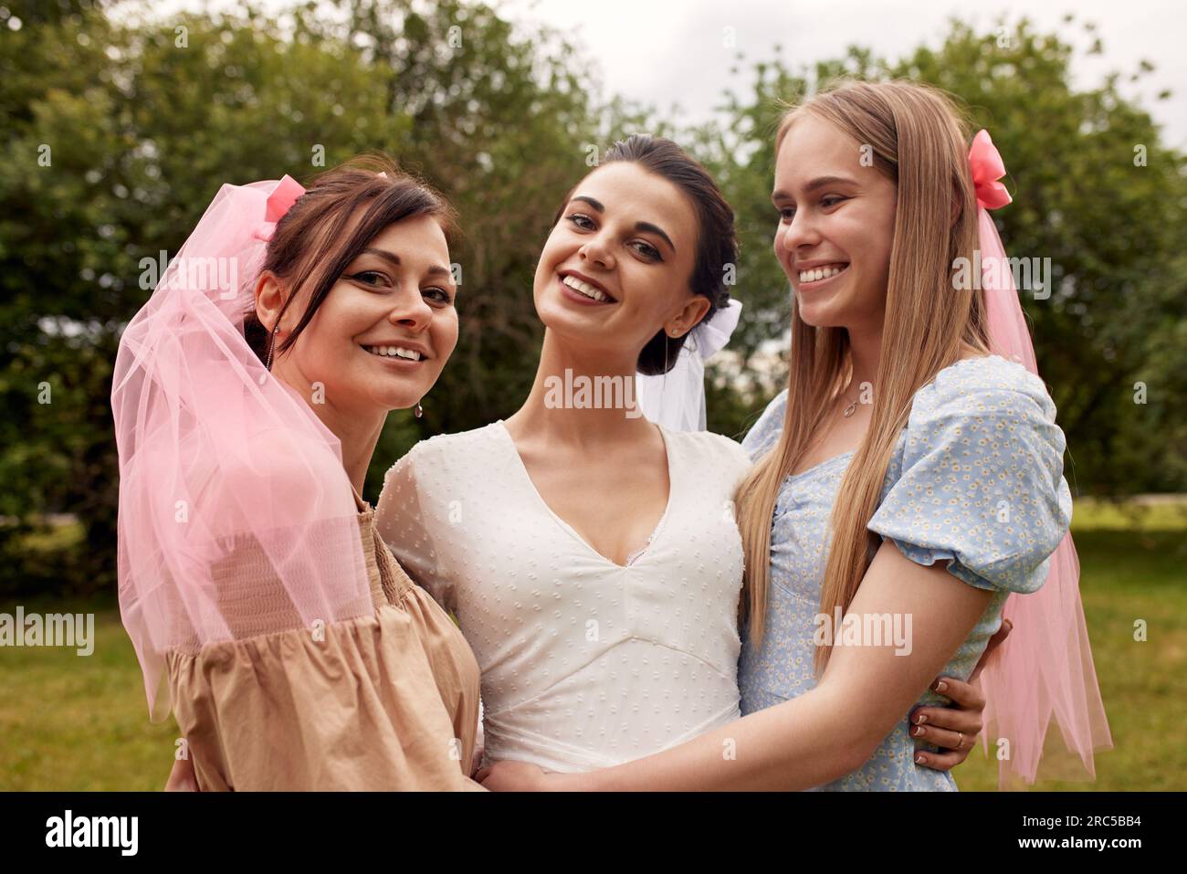 Heureuse mariée adorable dans une robe blanche avec un voile blanc et deux demoiselles d'honneur dans une robe bleue et beige avec voile rose sourire et câlin dans un parc d'été. Banque D'Images