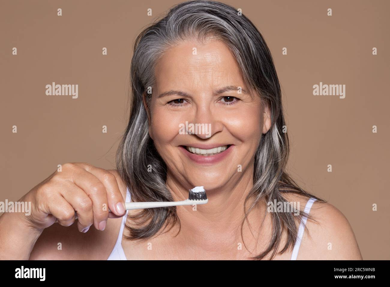 Joyeuse vieille dame caucasienne aux cheveux gris se brosse les dents, isolée sur fond brun, studio Banque D'Images