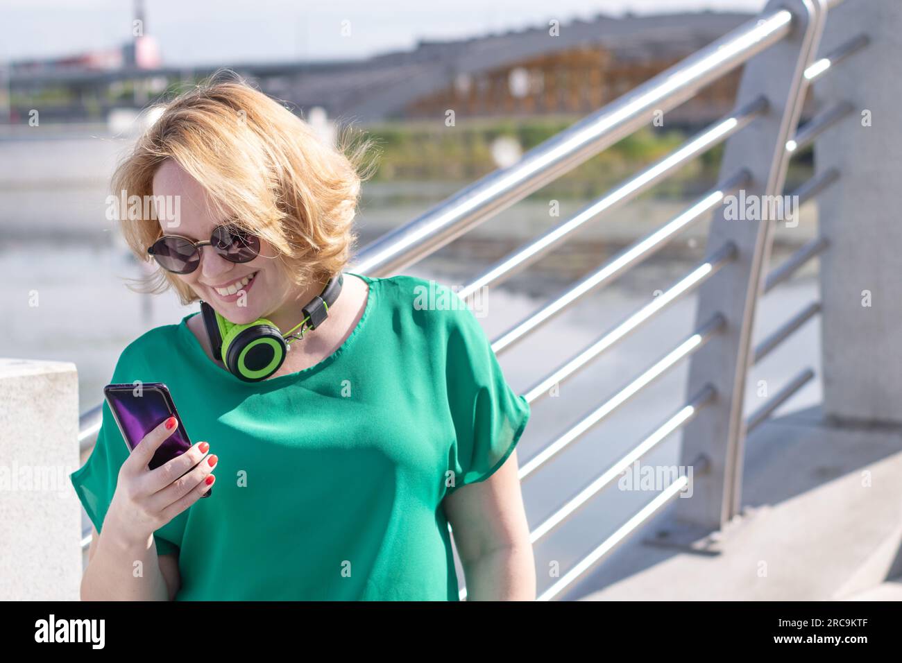 Une femme souriante avec des écouteurs tenant un téléphone dans la main étudie en ligne sur la rive. Heureuse fille étudiante dans des lunettes de soleil et des écouteurs en utilisant Banque D'Images