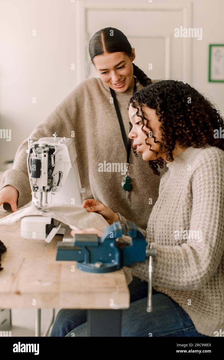 Enseignant souriant regardant une étudiante à l'aide d'une machine à coudre dans une classe d'art au lycée Banque D'Images