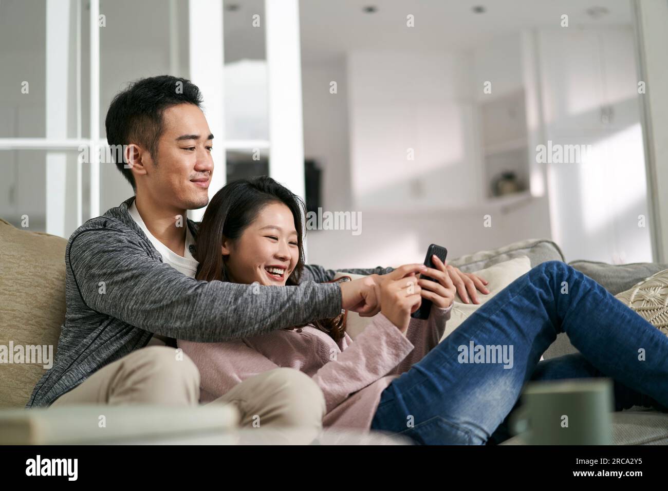 heureux jeune couple asiatique assis sur le canapé de la famille à la maison regardant des photos de téléphone portable ensemble Banque D'Images