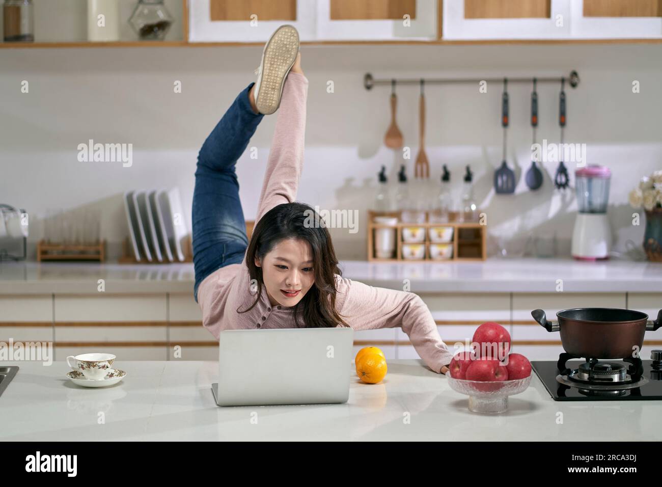 jeune femme d'affaires asiatique freelance étirant les jambes tout en travaillant dans la cuisine à la maison à l'aide d'un ordinateur portable Banque D'Images