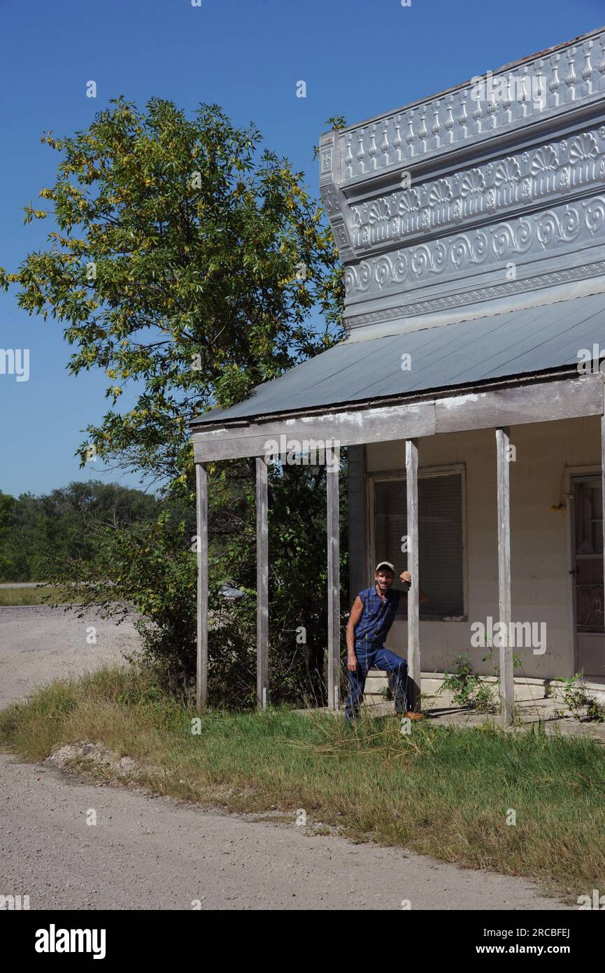 L'homme se penche contre le porche du vieux magasin de campagne sur la route de terre dans le Kansas. La façade du magasin est recouverte d'une boîte décorative. Banque D'Images