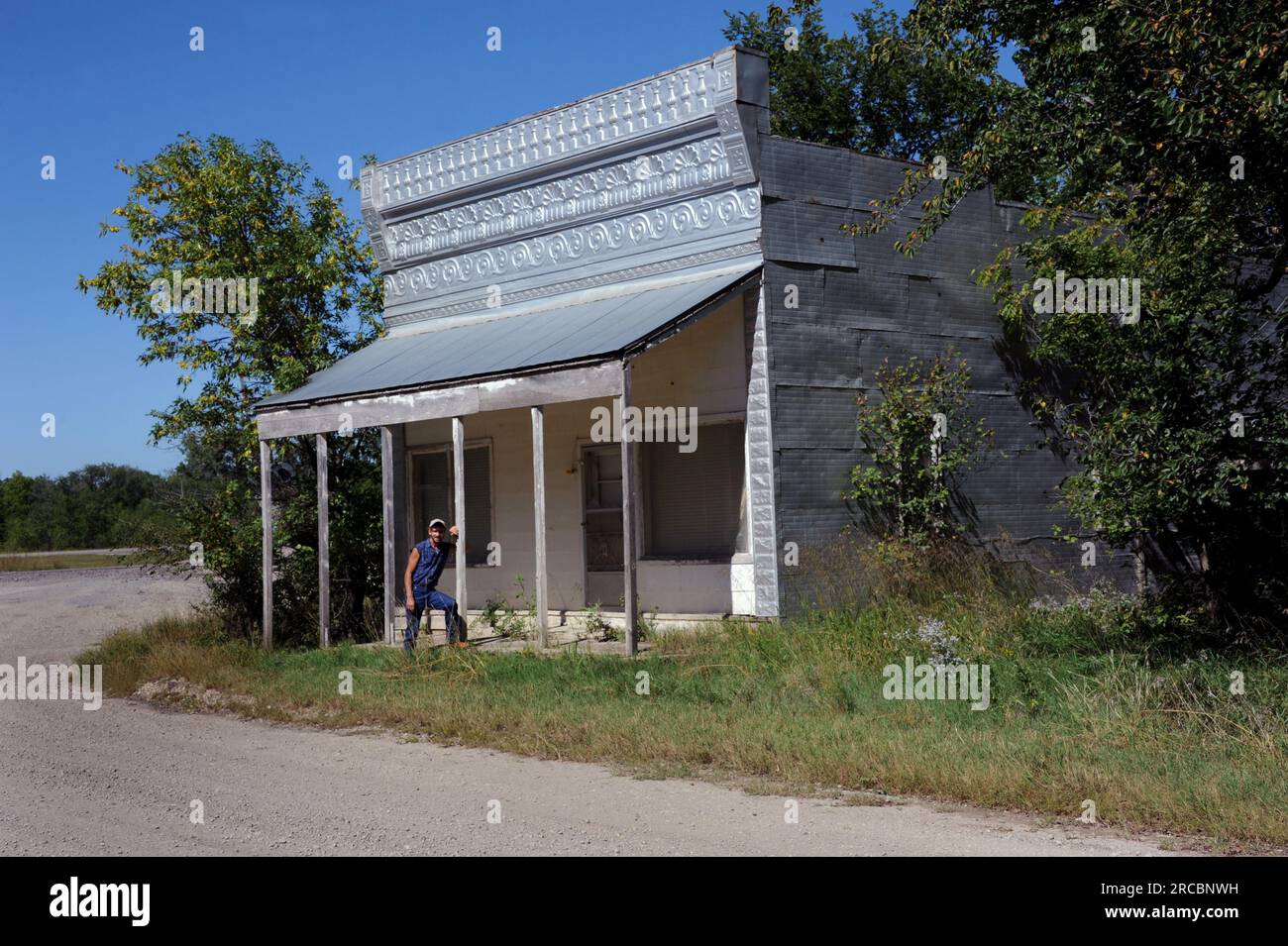 Des feuilles décoratives en étain couvrent le toit, le côté et l'avant du magasin. Le magasin abandonné est sur un chemin de terre au Kansas. L'homme pose par le poteau de la porte d'entrée. Banque D'Images
