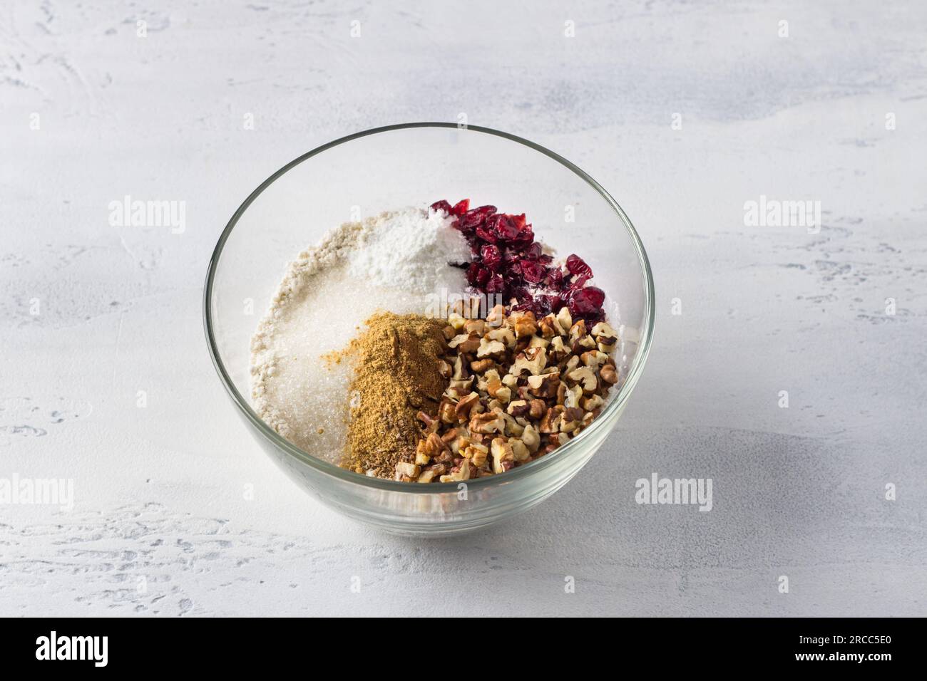 Bol en verre avec un mélange de farine, sucre, épices, noix et canneberges sur fond bleu clair. Cuisiner de délicieux gâteaux faits maison. Banque D'Images