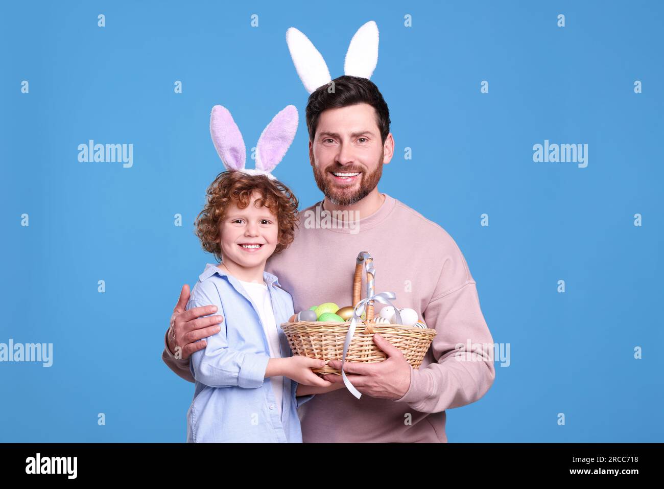 Heureux père et fils dans des bandeaux mignons d'oreilles de lapin tenant le panier de Pâques avec des œufs peints sur fond bleu clair Banque D'Images