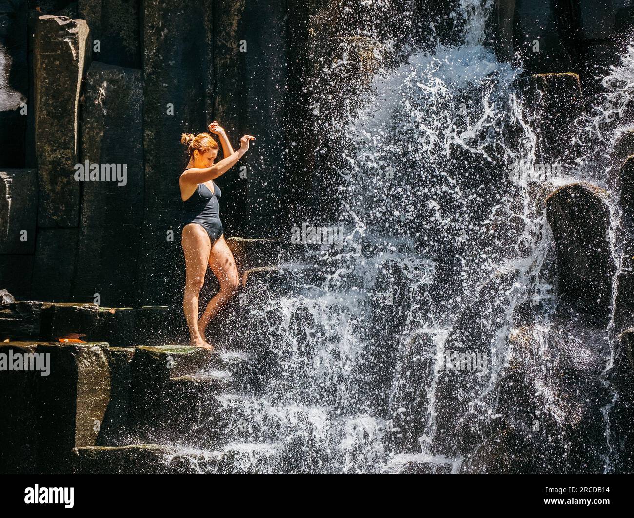 Femme caucasienne dans un maillot de bain noir rafraîchissant sous des ruisseaux d'eau tombant sur des cascades de pierre volcanique noire. Chutes d'eau de Rochester - Pooul Banque D'Images
