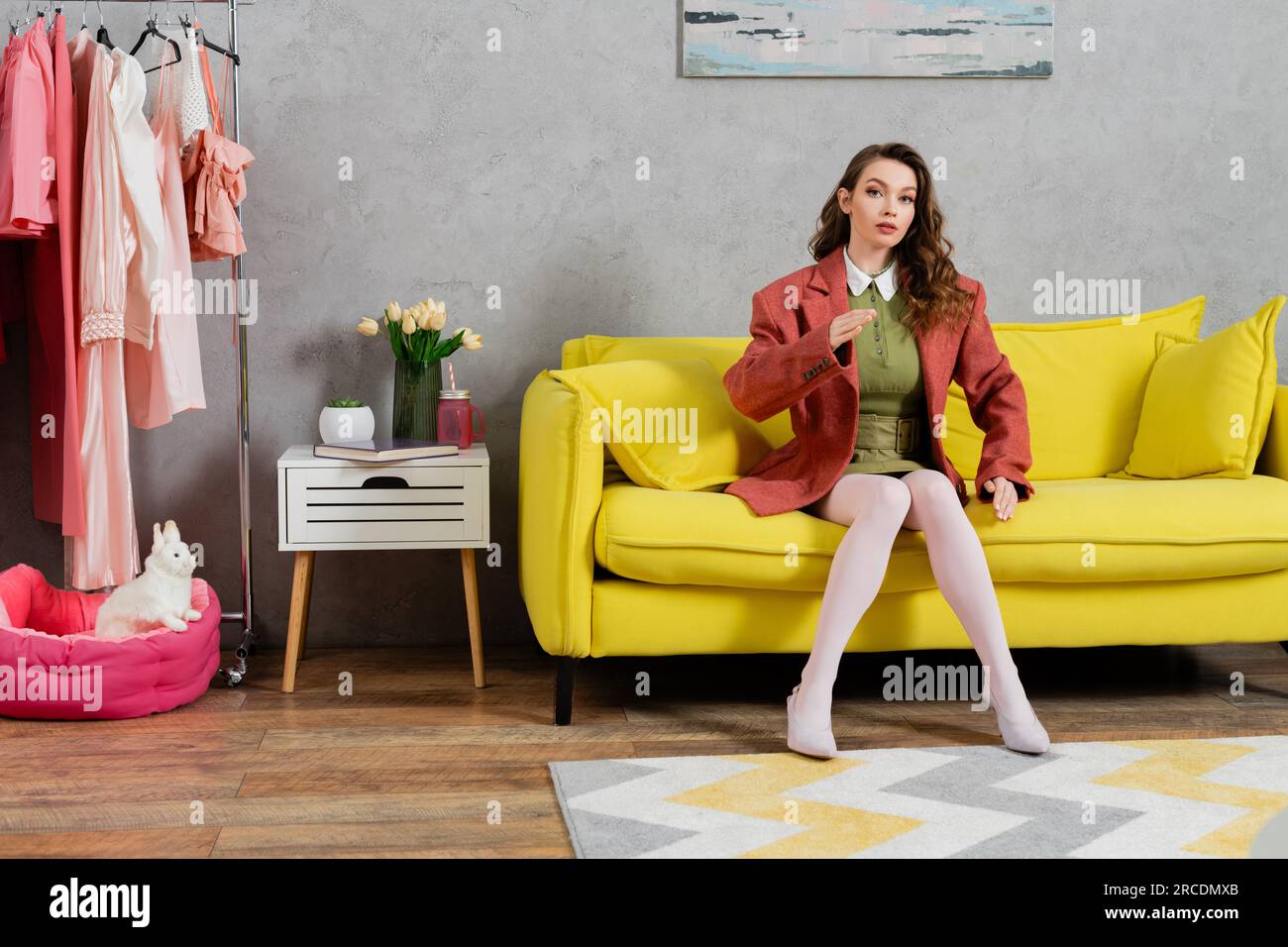 photographie de concept, posant comme une poupée, jeune femme bien habillée avec les cheveux ondulés assis sur le canapé jaune, gestuelle, intérieur de maison élégant, vase avec Banque D'Images