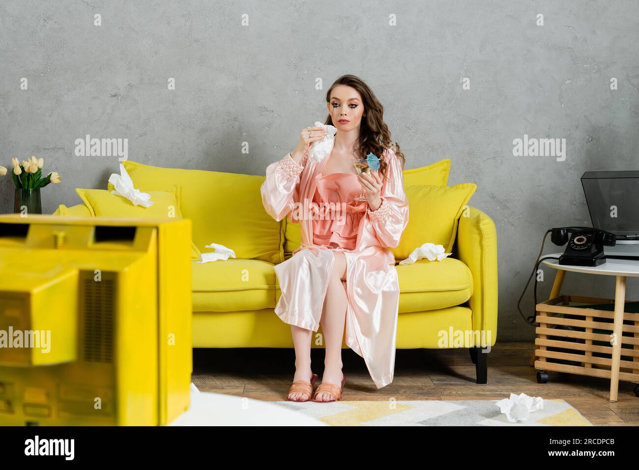 stress émotionnel, femme triste avec du mascara taché assis sur un canapé jaune, femme au foyer avec des yeux pleurant tenant une serviette et un cocktail, regardant la télévision, déprimant Banque D'Images