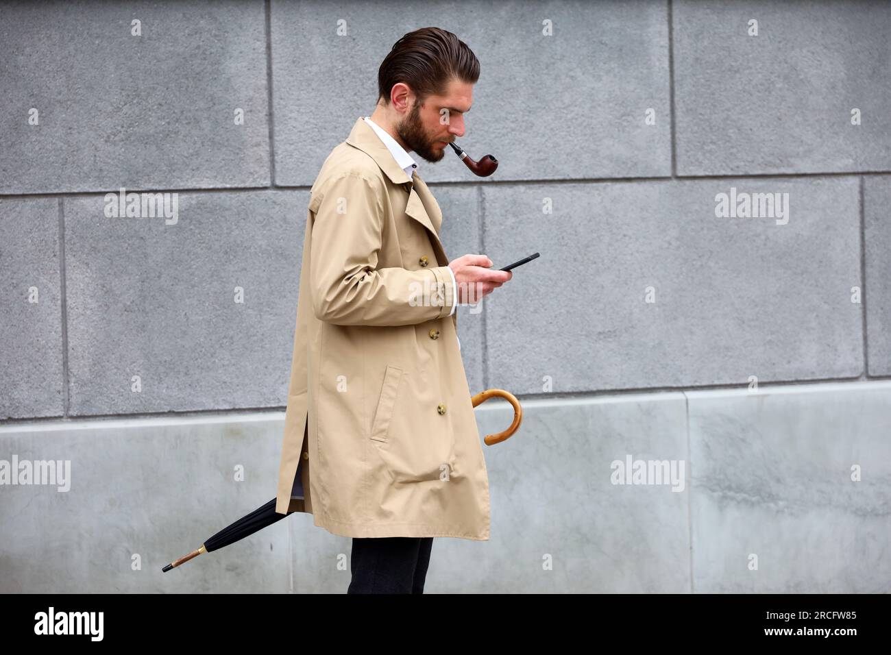 Homme avec une pipe à fumer portant un imperméable regardant l'écran du smartphone tout en se tenant dans la rue Banque D'Images