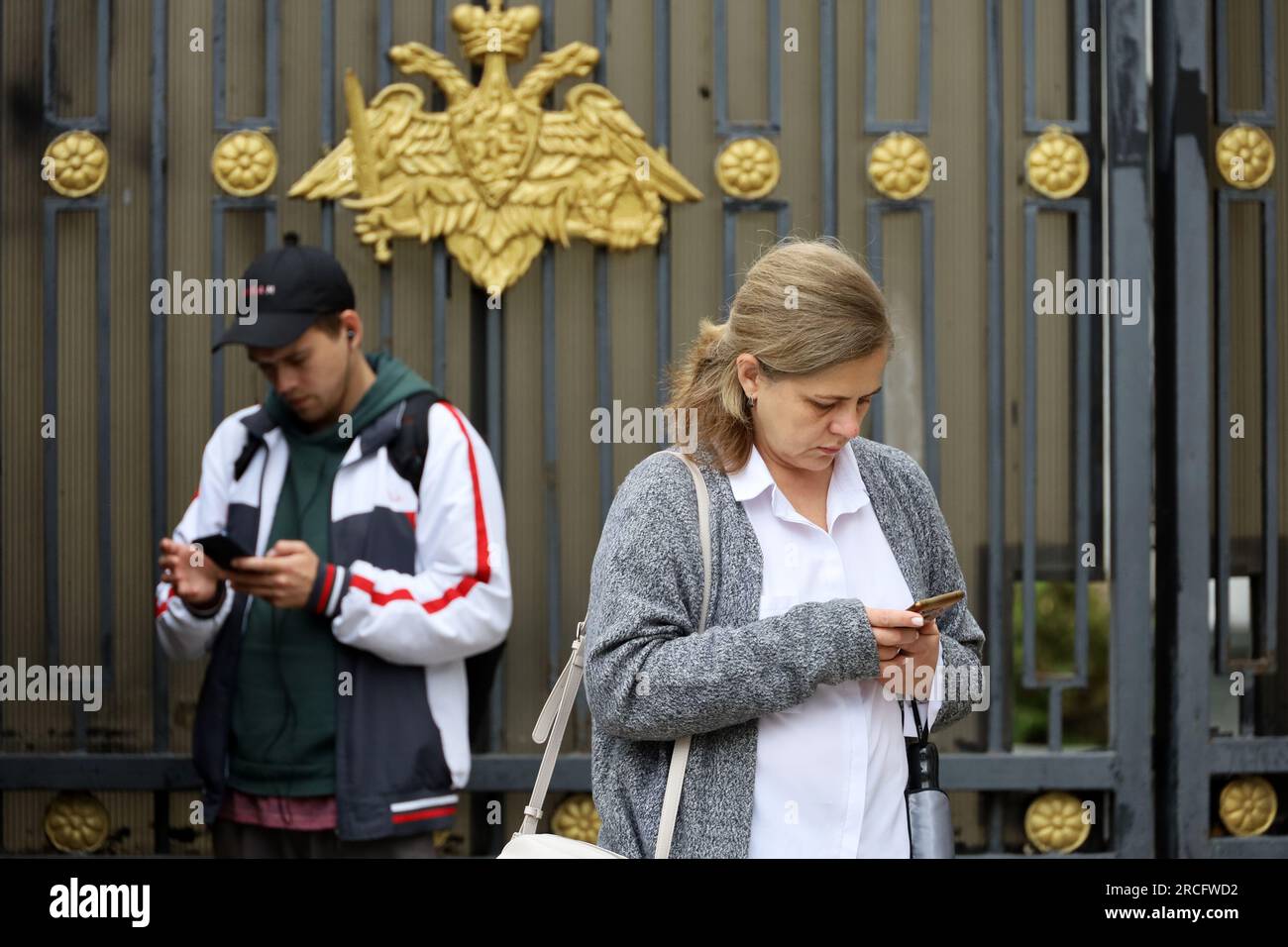 Inquiet femme et gars avec des smartphones debout sur la rue de la ville près de la clôture avec l'emblème officiel du ministère de la Défense de la Russie Banque D'Images
