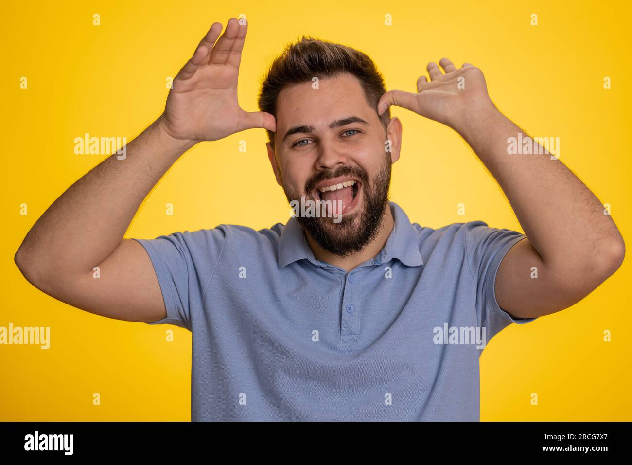 Comique drôle jeune homme caucasien faisant des expressions faciales stupides et ludiques et grimacant, trompant autour, montrant la langue. Beau gars adulte fou intimidant isolé sur fond jaune studio à l'intérieur Banque D'Images