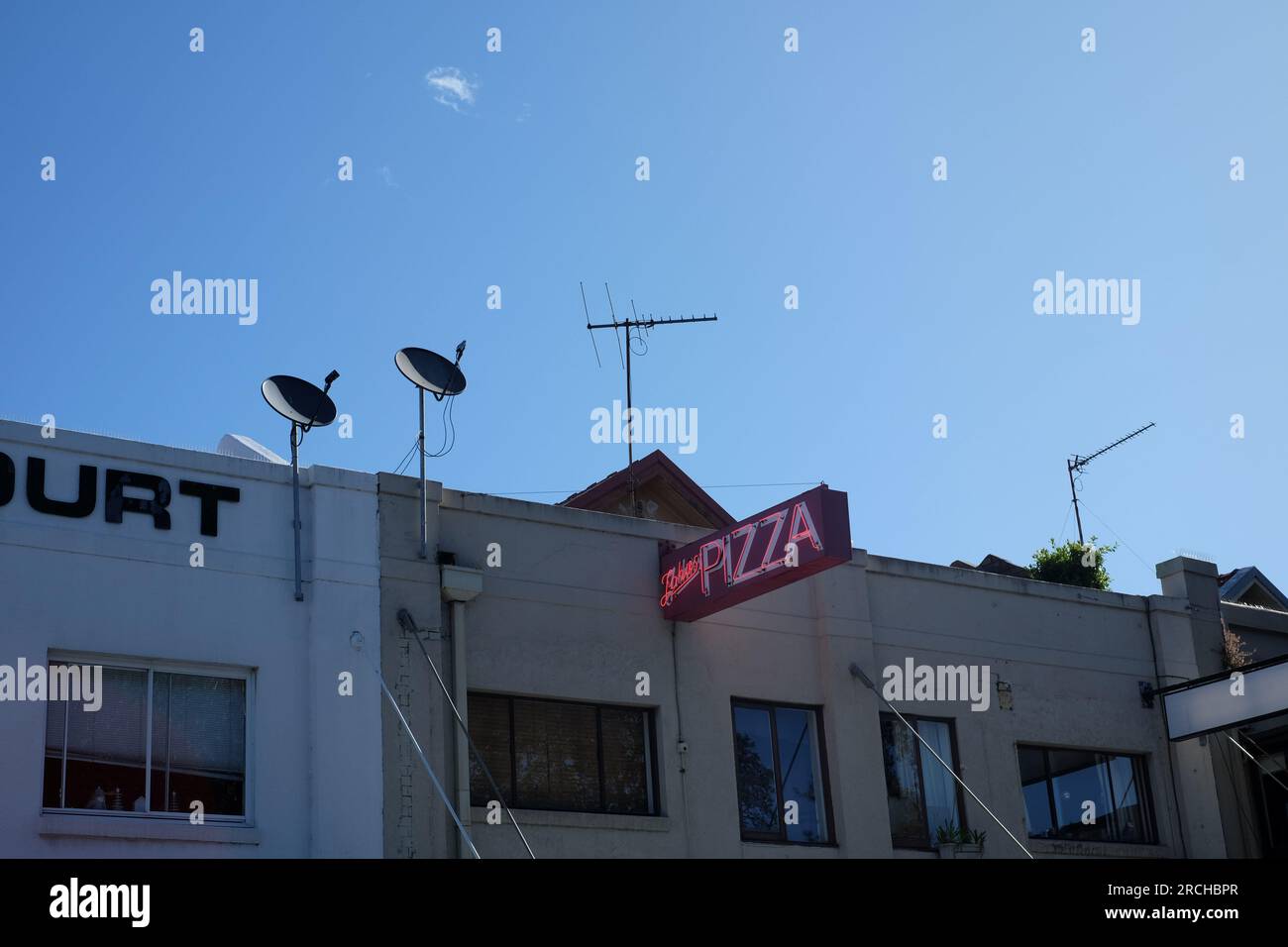 Un panneau de pizza rose et rouge au néon brille dans la lumière de l'après-midi, deux antennes paraboliques et des antennes de télévision montées sur le toit au-dessus d'une rangée de magasins Banque D'Images