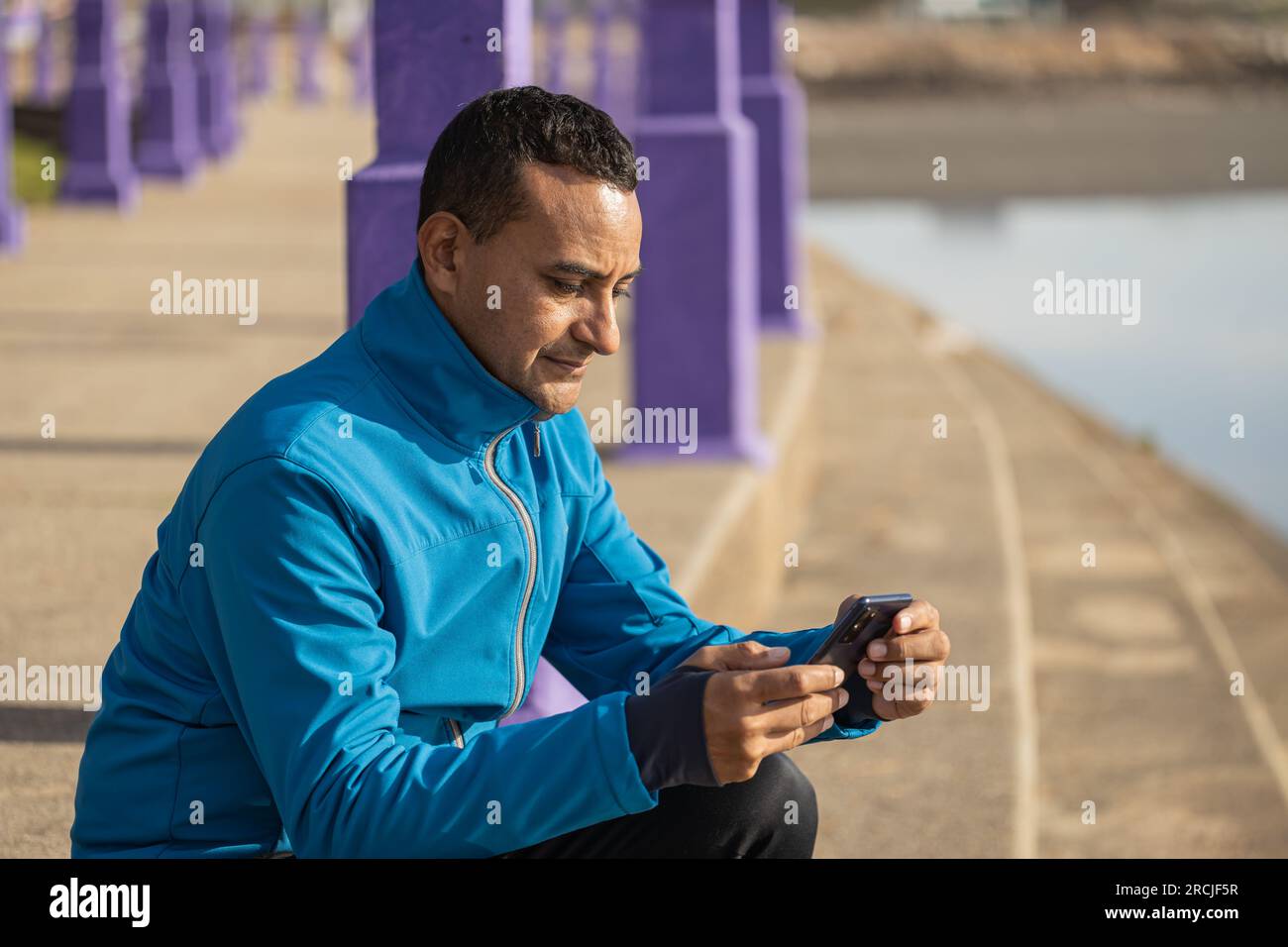 Plan moyen d'un jeune latino en vêtements de sport regardant son téléphone portable dans un parc public. Banque D'Images