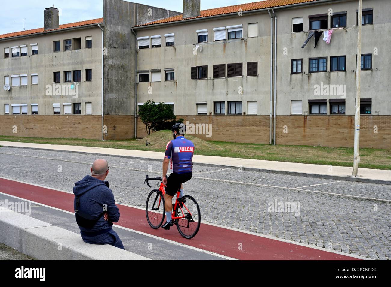 Homme assis sur le mur regardant un cycliste en vêtements de cyclisme passer avec bloc de vieux appartements derrière, Espinho, nord du Portugal Banque D'Images