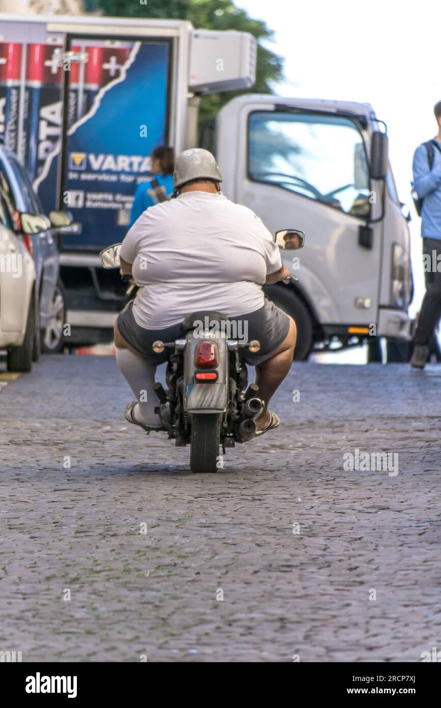 Une personne sur un petit scooter à moteur traverse une rue pavée, passant des camions et des piétons dans un cadre urbain - la Valette, Malte. Banque D'Images
