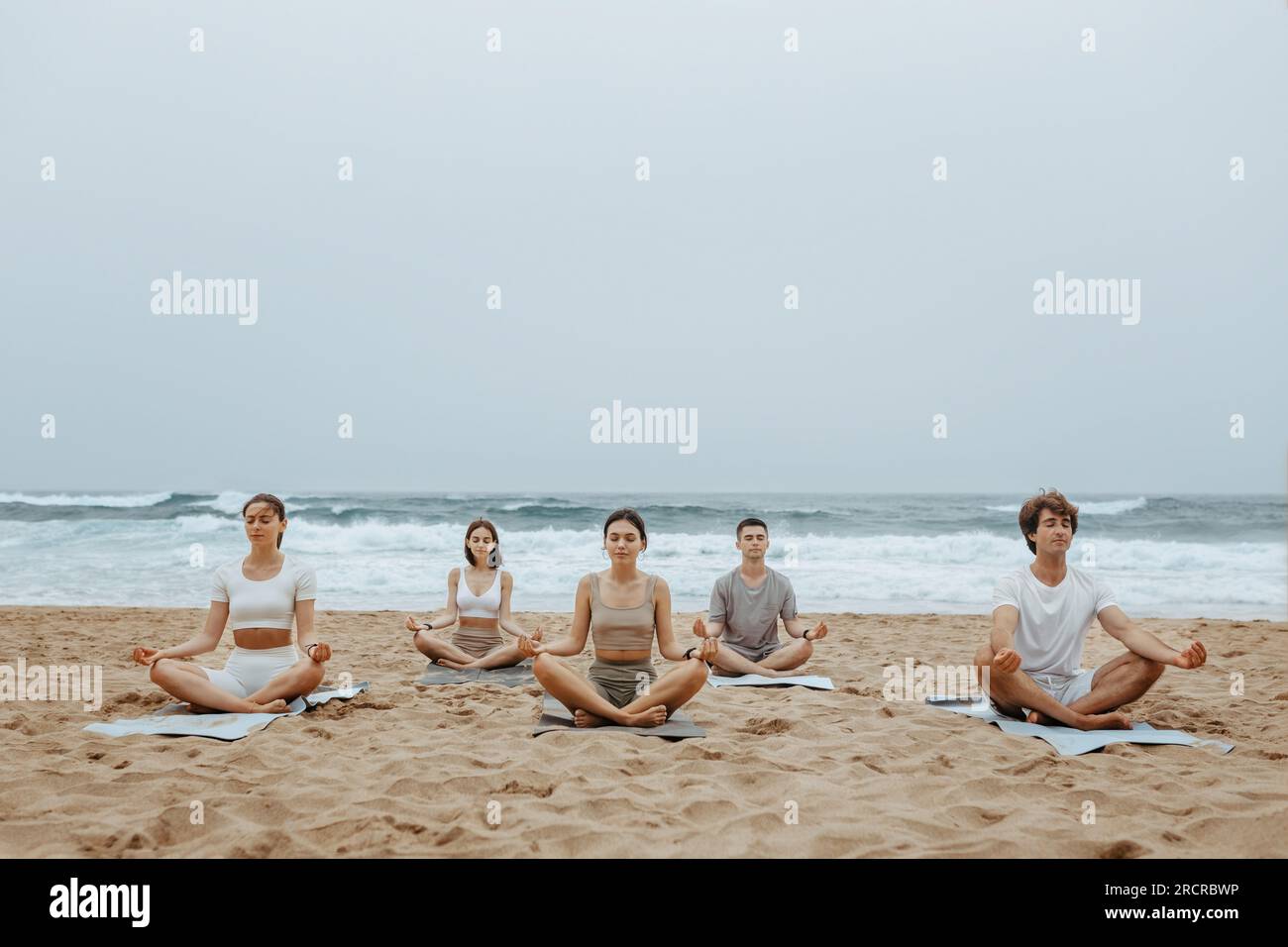 Yoga de plage pour les jeunes. Groupe de femmes et d'hommes méditant à la plage sur la rive de l'océan, assis sur des nattes sur le sable, espace libre Banque D'Images
