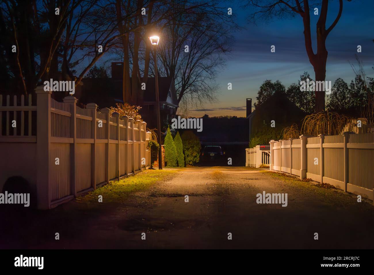 Découvrez la magie du crépuscule tandis qu'un lampadaire vous guide devant de pittoresques clôtures blanches dans ce village de Nouvelle-Angleterre. Chaque détail murmure une histoire. Banque D'Images