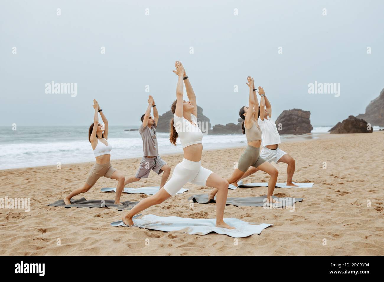 Cours de yoga sur la rive de l'océan. Groupe de jeunes faisant la pose de guerrier, méditant et faisant de l'exercice sur la plage Banque D'Images