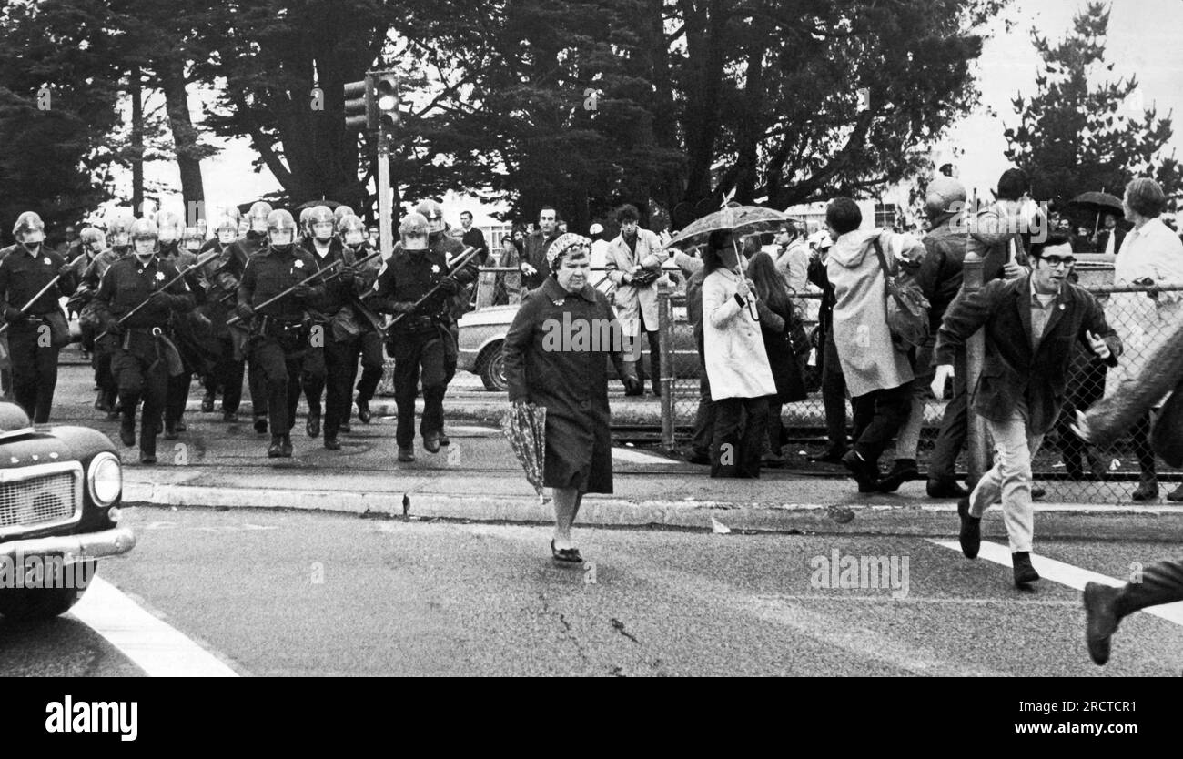 San Francisco, Californie : 9 décembre 1968 une dame de parapluie qui traverse la 19e Avenue, ignorant complètement que l'escadron de police en équipement anti-émeute double chronométrage derrière elle à la poursuite des manifestants fuyant l'Université d'État de San Francisco. Banque D'Images