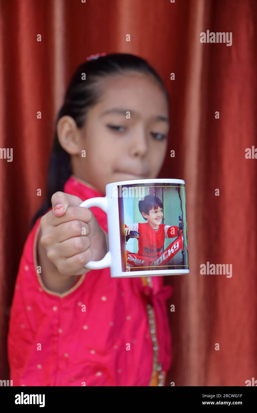 Une petite jolie fille indienne dans diverses poses avec sa tasse à café qui a sa propre photo imprimée dessus. robe rouge. Banque D'Images