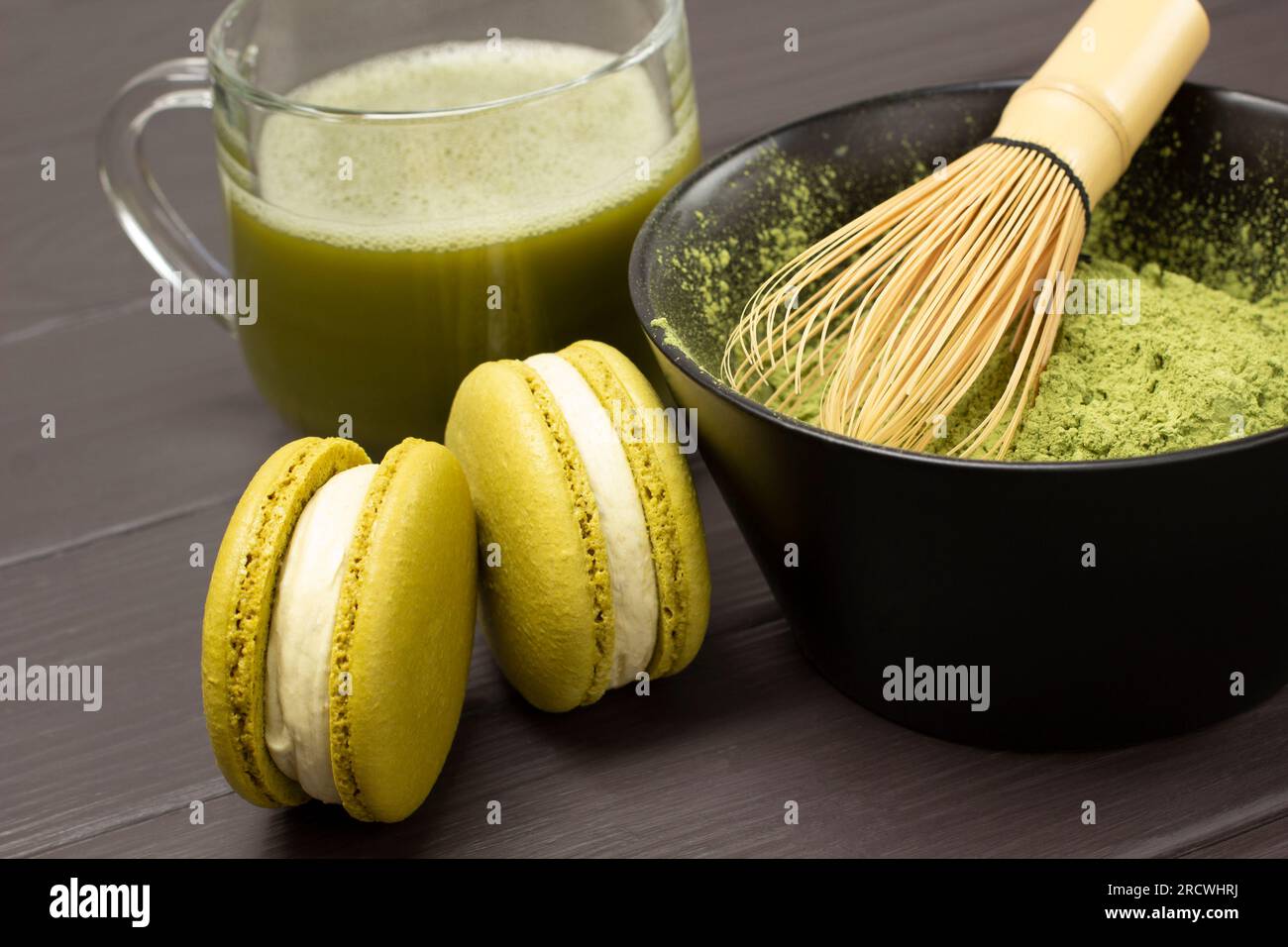 Fouetter en bambou dans un bol avec de la poudre de matcha. Deux gâteaux de macaroni et une tasse de thé vert matcha. Gros plan. Fond en bois foncé Banque D'Images
