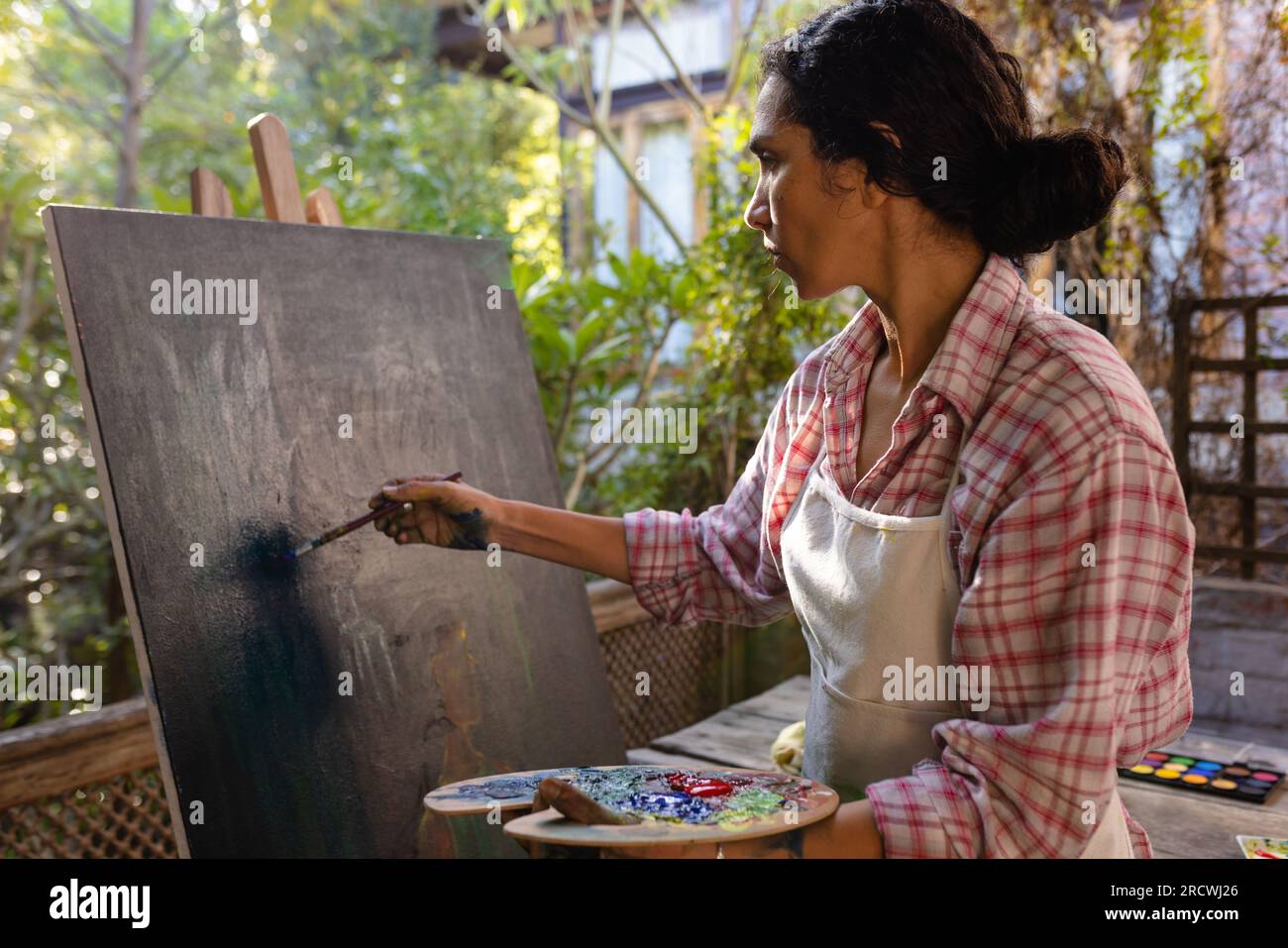 Femme biracial portant la peinture de tablier sur le chevalet en utilisant la brosse et la palette de couleurs dans le jardin Banque D'Images