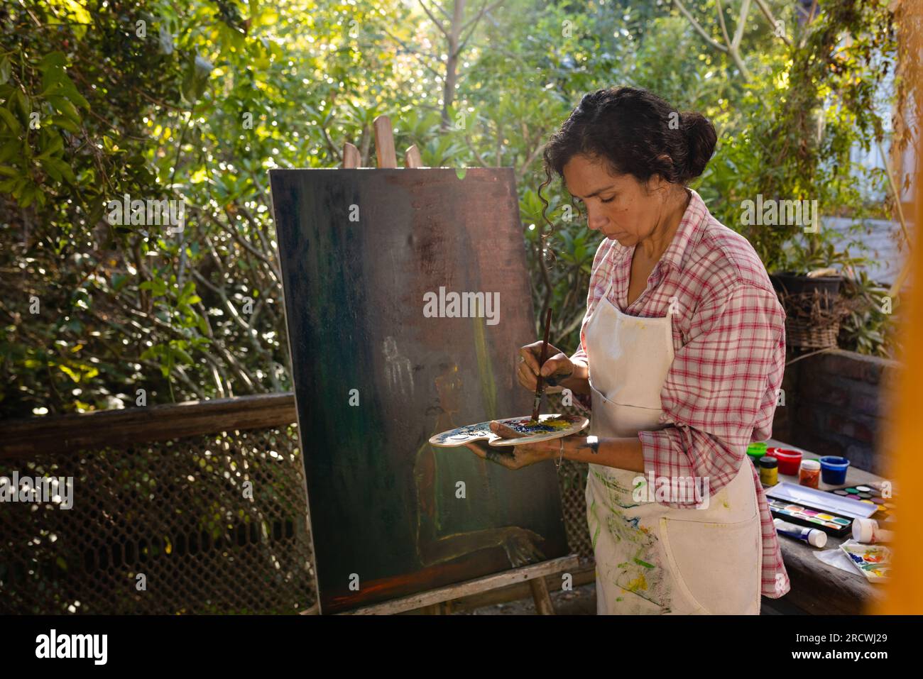 Femme biracial portant la peinture de tablier sur le chevalet en utilisant la brosse et la palette de couleurs dans le jardin Banque D'Images