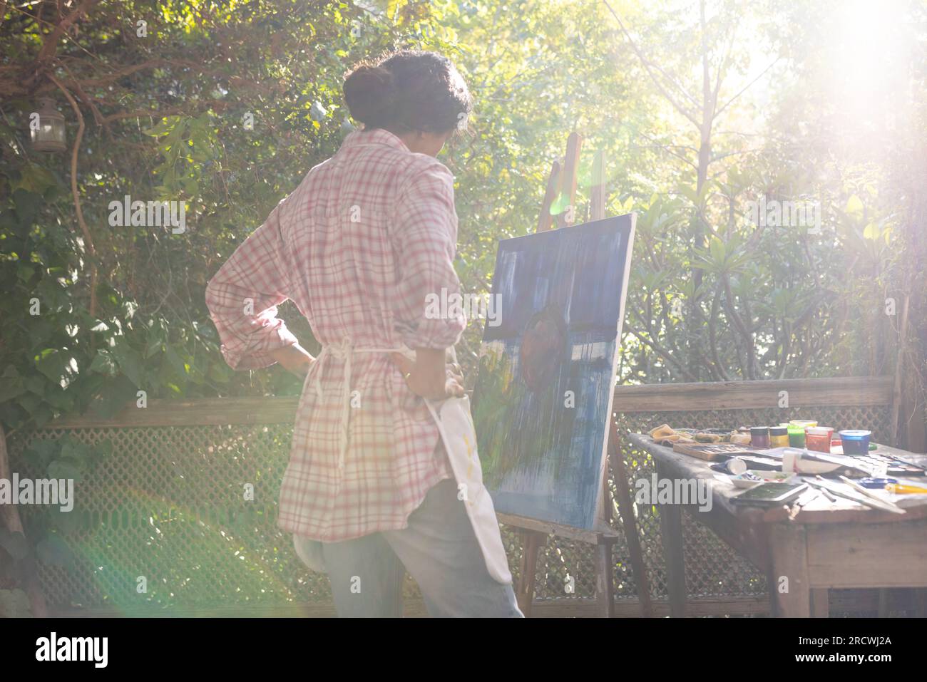 Femme biraciale portant un tablier regardant la peinture sur chevalet dans le jardin Banque D'Images