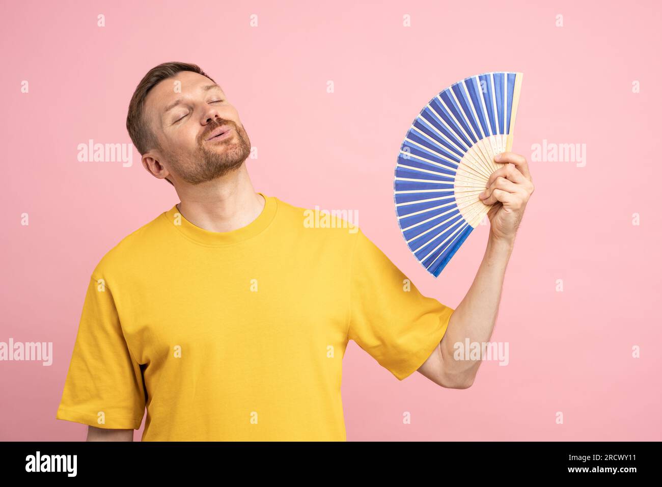 Homme surchauffé utilisant le ventilateur de papier souffrent de la chaleur, profitant de l'air frais isolé sur le mur rose studio Banque D'Images