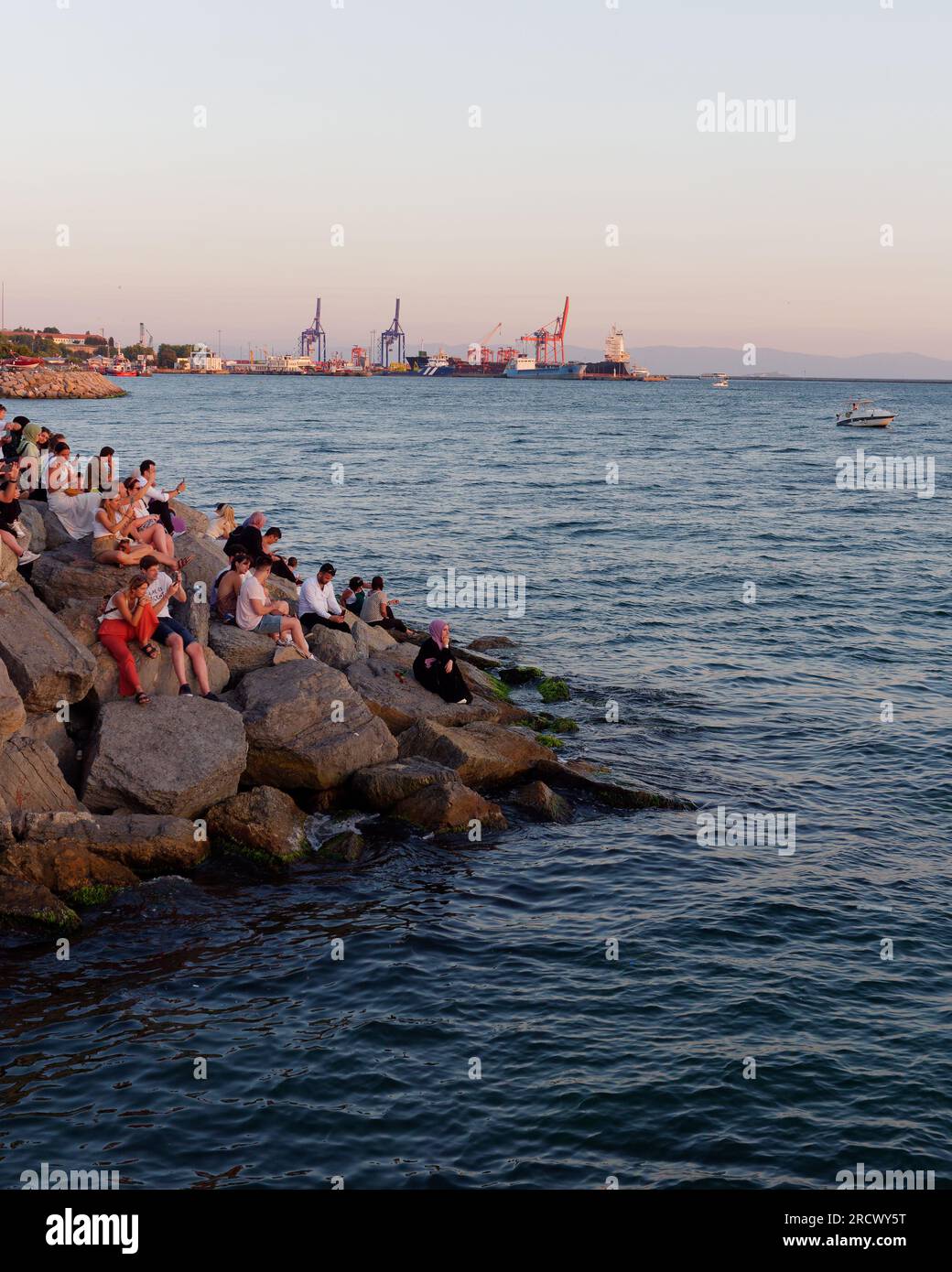 Les gens s'assoient sur les rochers et profitent du coucher de soleil d'été sur la mer du Bosphore depuis Uskudar, Istanbul, Turquie. Grues à Kadikoy en arrière-plan Banque D'Images