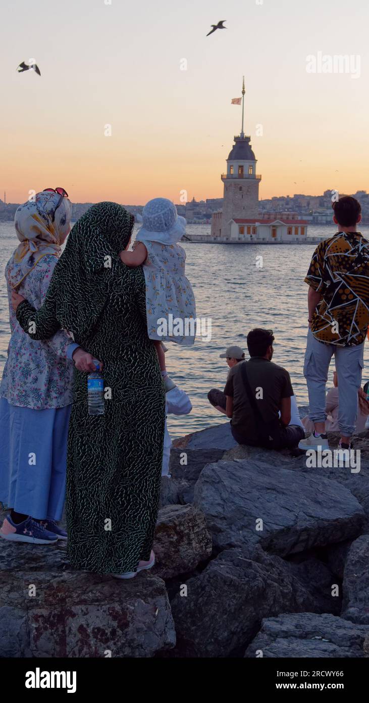 Deux femmes portant un enfant se tiennent debout sur des rochers profitant du coucher de soleil de l'été sur la mer du Bosphore et la Tour Maidens d'Uskudar, Istanbul, Turquie Banque D'Images
