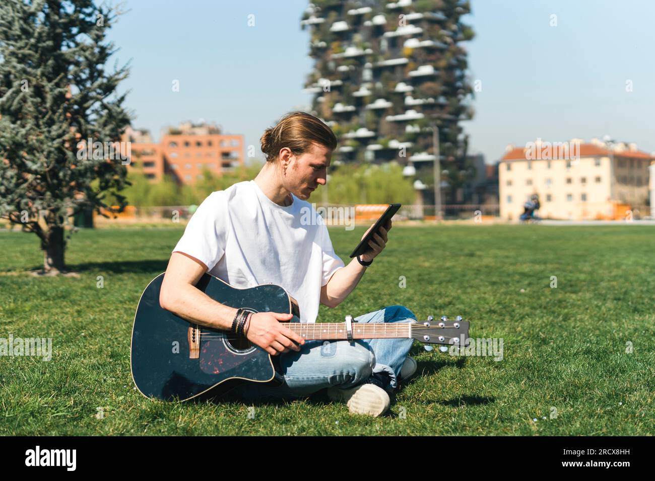 homme regardant le dispositif de tablette tenant la guitare dans un jardin urbain Banque D'Images