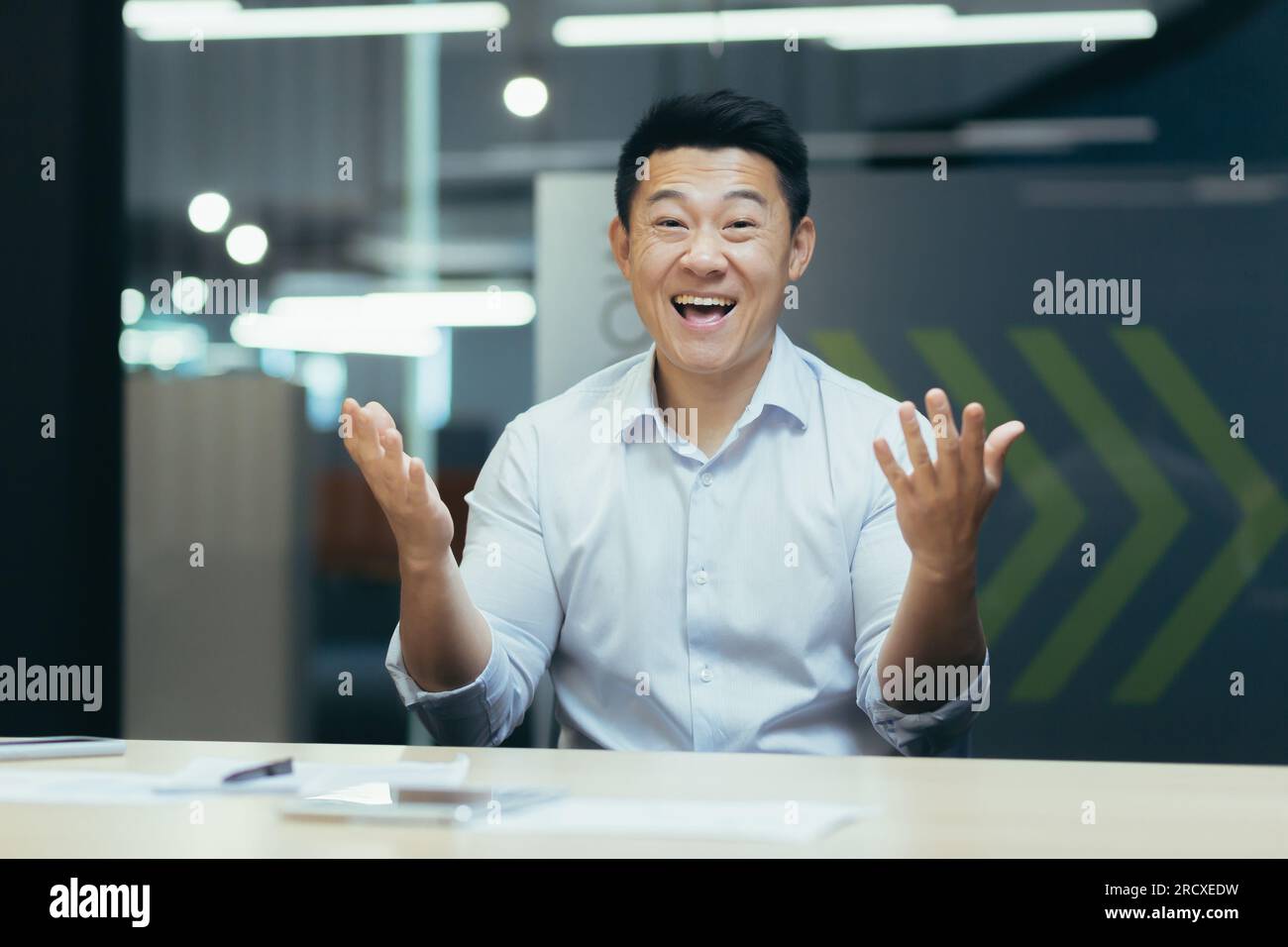 Portrait d'un jeune homme asiatique heureux assis à un bureau devant une caméra et parlant lors d'un appel vidéo. Faisant des gestes avec ses mains, souriant à la caméra. Banque D'Images