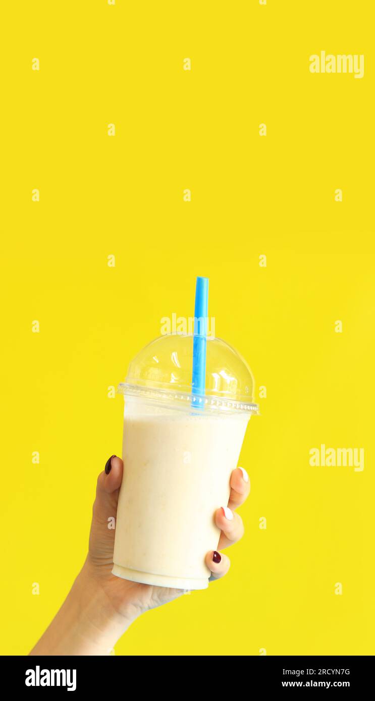 Smoothie banane avec tasse jetable en plastique. Milkshake banane dans la main d'une fille sur un fond jaune vif. Espace de copie Banque D'Images