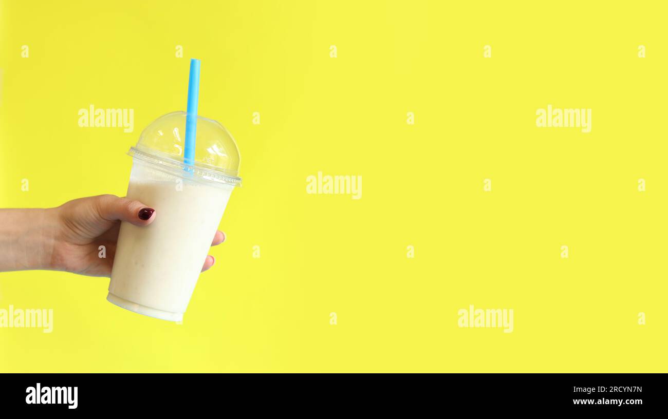 Smoothie banane avec tasse jetable en plastique. Milkshake banane dans la main d'une fille sur un fond jaune vif. Espace de copie Banque D'Images