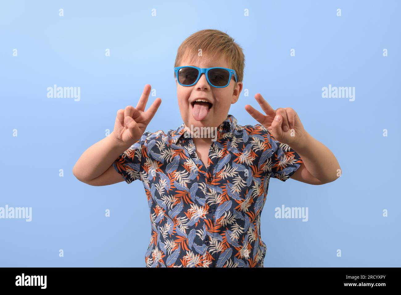 Portrait d'un adolescent dans une chemise hawaïenne et des lunettes de soleil bleues. Les mains levées se réjouissent en regardant la caméra. Banque D'Images