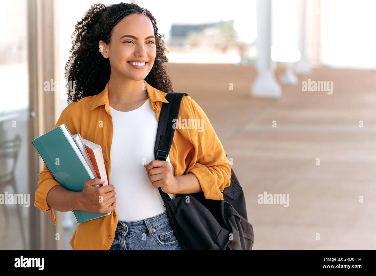 Photo d'une belle étudiante brésilienne ou hispanique aux cheveux bouclés positive, avec un sac à dos, tenir des livres et des cahiers dans sa main, marcher près du campus universitaire, regarder loin et sourire heureux Banque D'Images