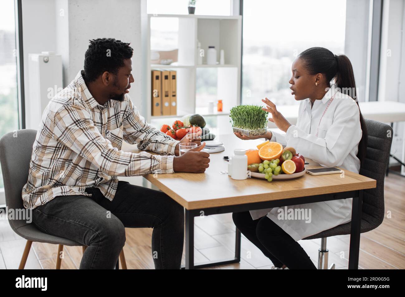 Femme attrayante en blouse de laboratoire donnant la consultation à l'homme adulte sur l'utilisation de pousses de légumes de salade dans l'alimentation quotidienne. Dame multiethnique sérieuse relatant les composants de soutien de la santé des microgreens. Banque D'Images
