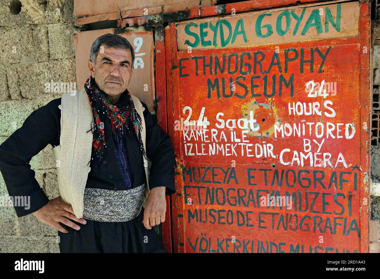 Seyda Goyan est vu en vêtements nationaux kurdes devant la porte intéressante de son musée. Sur la porte, le nom du musée est écrit en 7 langues différentes pour les visiteurs. Le folkloriste Seyda Goyan, qui a écrit 6 livres sur le folklore kurde, a fondé le « Musée ethnographique kurde Seyda Goyan », où environ un millier d'œuvres ethnographiques sont stockées et exposées de ses propres moyens. Seyda Goyan, qui n’a pas pu trouver de lieu d’exposition pour les œuvres qu’il a recueillies en trois ans, a fait de sa maison historique de 2 pièces un musée dans le quartier d’Uludere, dans la province de Sirnak en Turquie. Banque D'Images