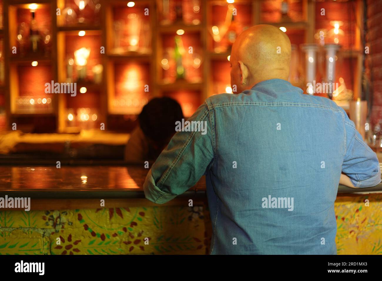 Jeune et flamboyant beau gars chauve tiré de derrière sur le comptoir du bar. Homme attrayant appréciant dans l'intérieur du club de ville à la boîte de nuit. Commander de l'alcool. Banque D'Images