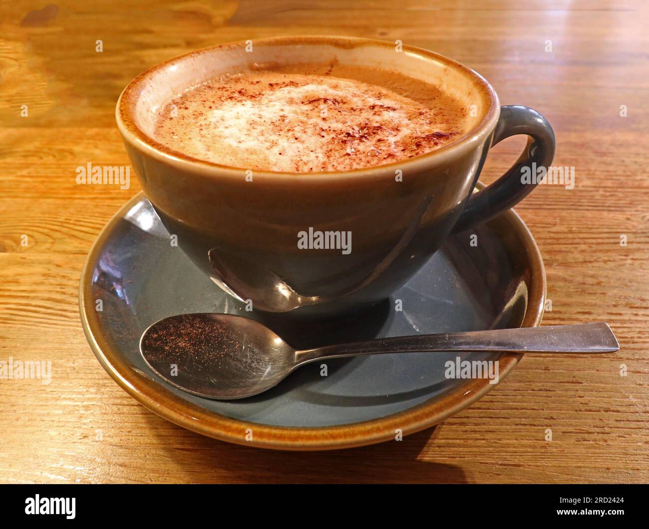 Café cappuccino servi dans une tasse en céramique avec soucoupe et cuillère sur une table en bois, photographié dans un cadre de café communautaire Banque D'Images