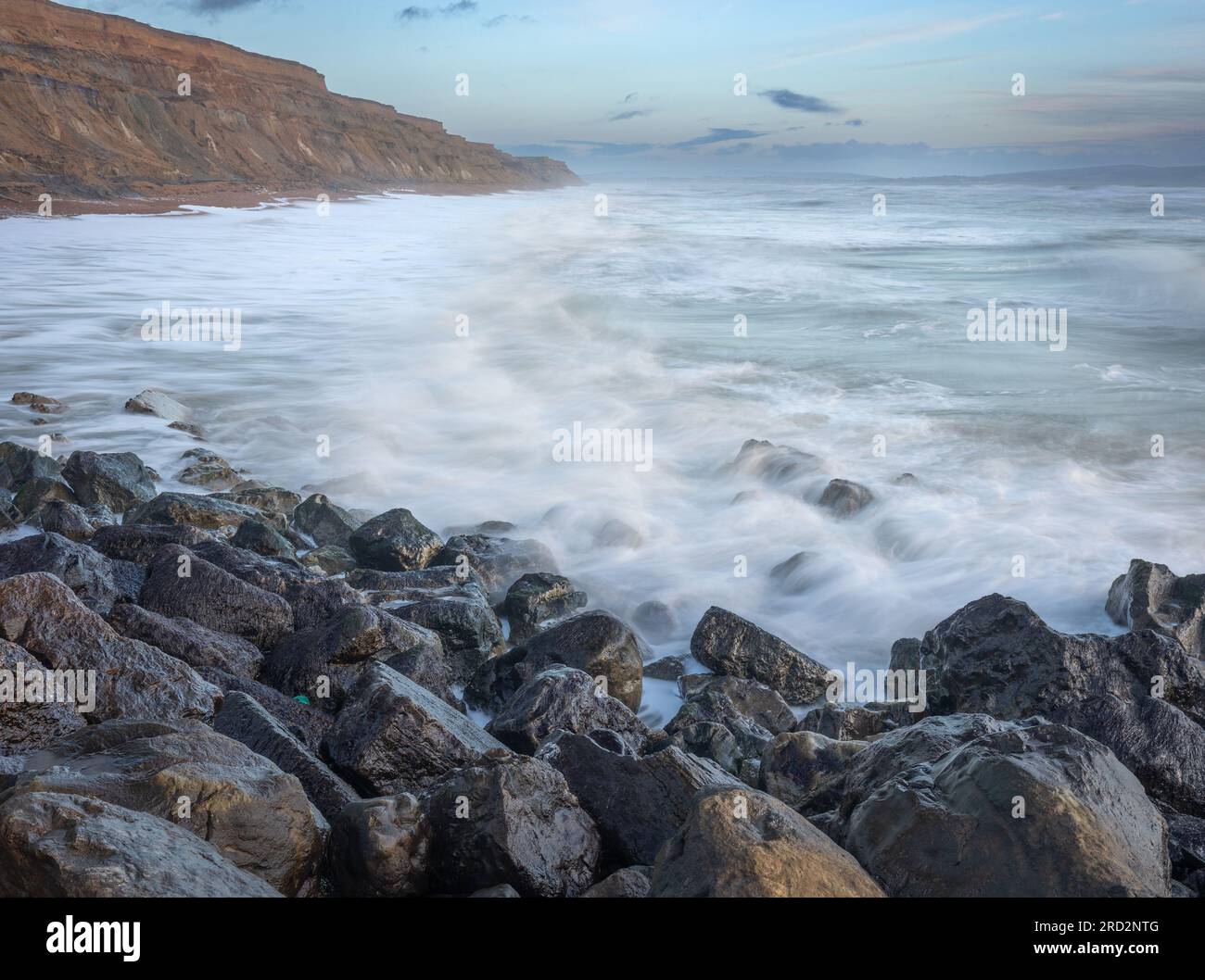 Une scène de paysage marin tôt le matin avec des rochers brillants au premier plan, des vagues floues sur les rochers et de hautes falaises de pierre de sable allant au loin Banque D'Images