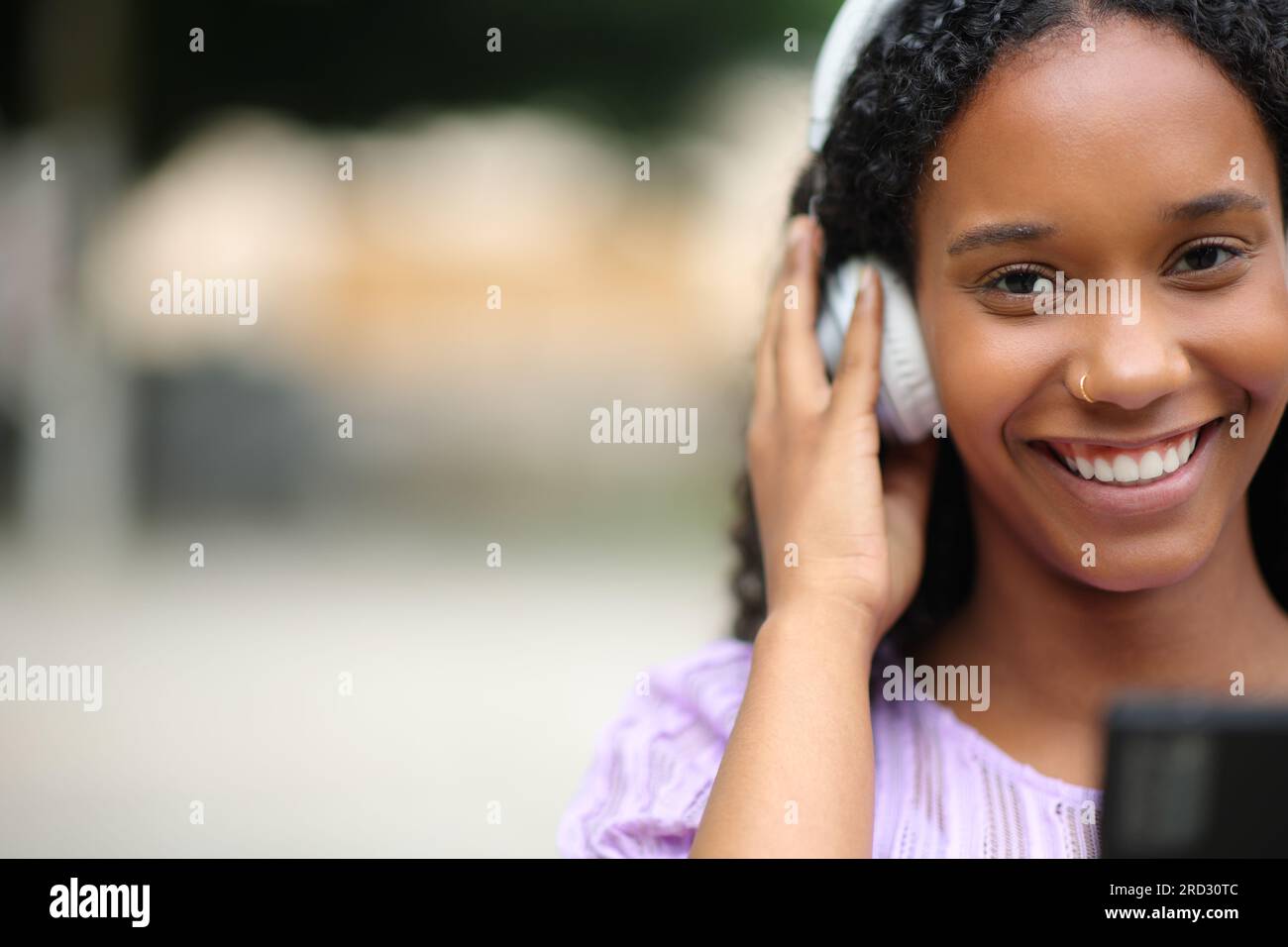 Vue de face d'une femme noire portant un casque et tenant le téléphone vous regardant écouter de la musique dans la rue Banque D'Images