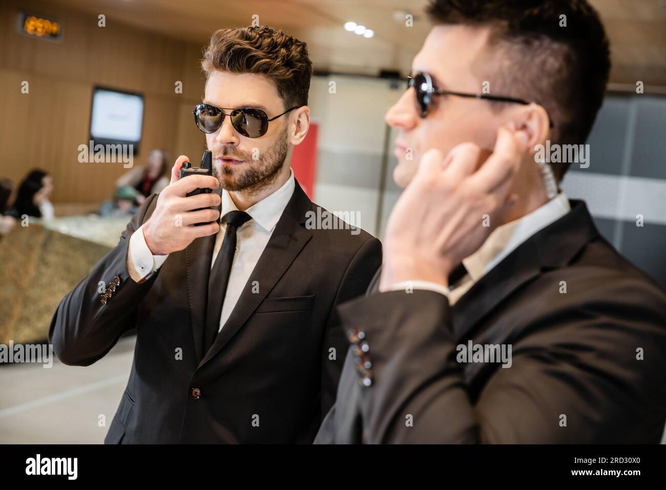 service bodyguard, sécurité privée, bel homme dans les lunettes de soleil et vêtements formels communiquant avec son partenaire de travail par talkie-walkie, sécurité de l'hôtel Banque D'Images