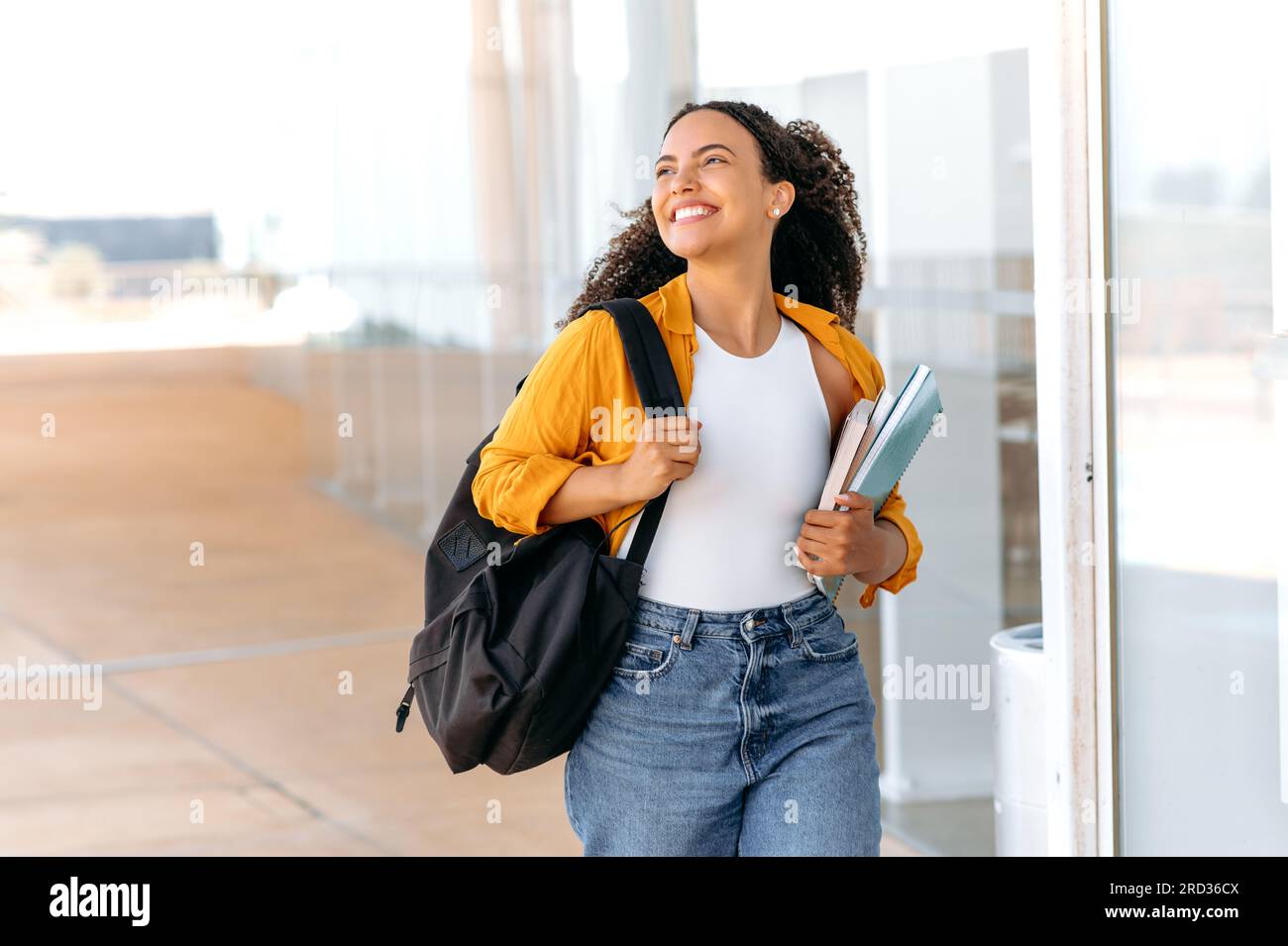 Heureuse belle étudiante brésilienne ou hispanique aux cheveux bouclés, avec un sac à dos, tenir des livres et des cahiers dans sa main, marcher près du campus universitaire, regarder loin et sourire, a terminé la journée d'école Banque D'Images