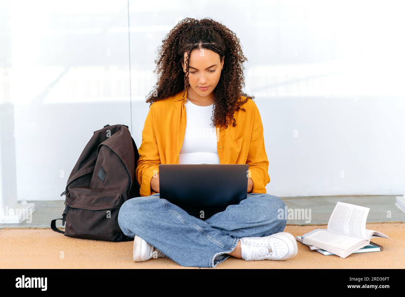Belle étudiante brésilienne ou hispanique aux cheveux bouclés focalisée et élégante, asseyez-vous près du campus universitaire, en utilisant un ordinateur portable, se préparer à l'examen, faire ses devoirs, tâche de projet, sourire Banque D'Images