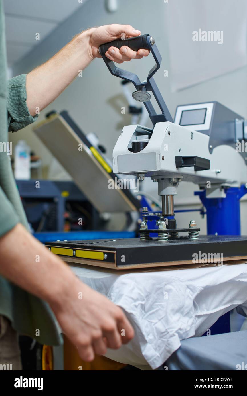 Vue recadrée d'un jeune artisan travaillant avec une machine de sérigraphie et des vêtements dans un studio d'impression flou à l'arrière-plan, une petite entreprise axée sur le client Banque D'Images